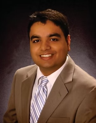 The image shows a man in a beige suit and striped tie, smiling against a dark background.