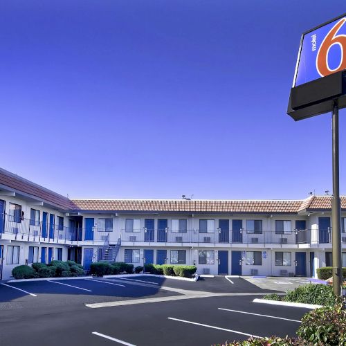 A motel with a two-story building, blue doors, and a parking area. A sign with the number 6 is prominently displayed in the foreground.