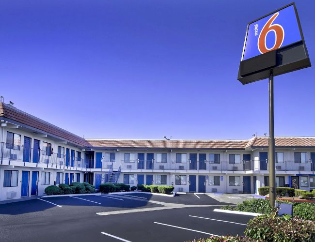 A motel with a two-story building, blue doors, and a parking area. A sign with the number 6 is prominently displayed in the foreground.