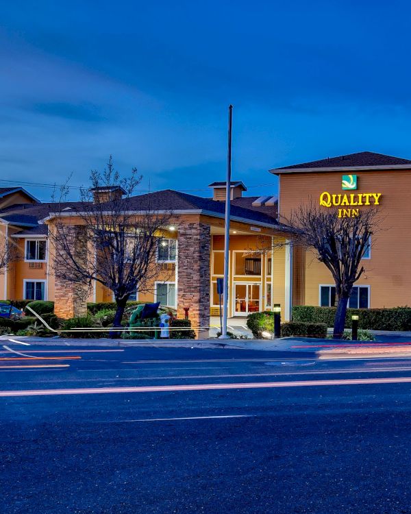 The image shows a Quality Inn hotel building with a well-lit exterior at dusk, cars parked in front, and light trails on the street.