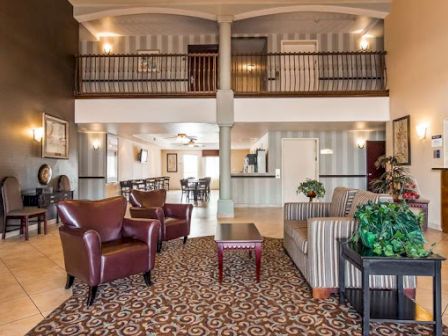 A cozy hotel lobby with seating area, plants, and a reception desk. There's a second-floor balcony overlooking the space.