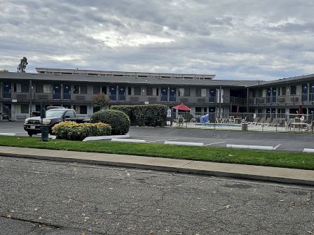 A two-story motel with a parking lot and a small pool area, surrounded by cars and bushes under a cloudy sky.