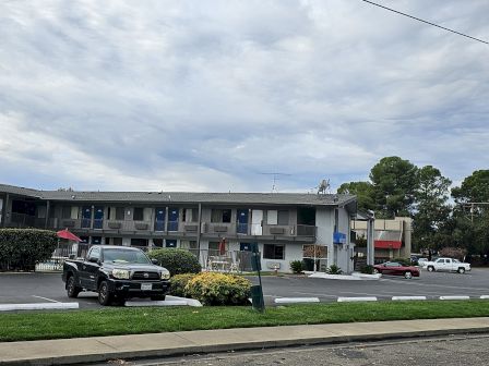 A Motel 6 building with a Denny's sign nearby, a parking lot, and a few vehicles are visible under a cloudy sky.