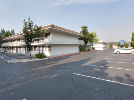 A two-story motel building with a parking lot and parked cars, surrounded by trees and clear sky.