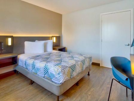 A modern hotel room with a bed, geometric bedspread, desk, chair, and wall-mounted TV, featuring wood flooring and minimal decor.