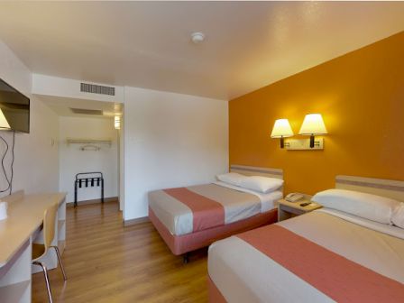 The image shows a hotel room with two beds, a TV, a desk, and warm lighting on an orange accent wall.