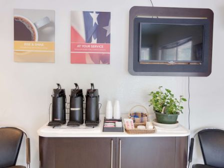 A coffee station with dispensers, cups, condiments, wall art, a TV, and plants, flanked by two chairs.