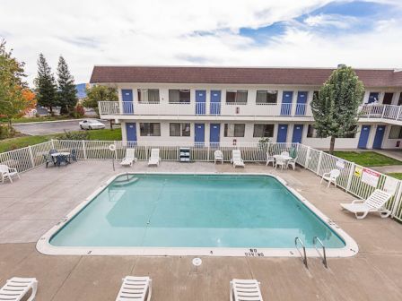 A motel with a swimming pool surrounded by loungers and trees, featuring a two-story building with blue doors and balconies.