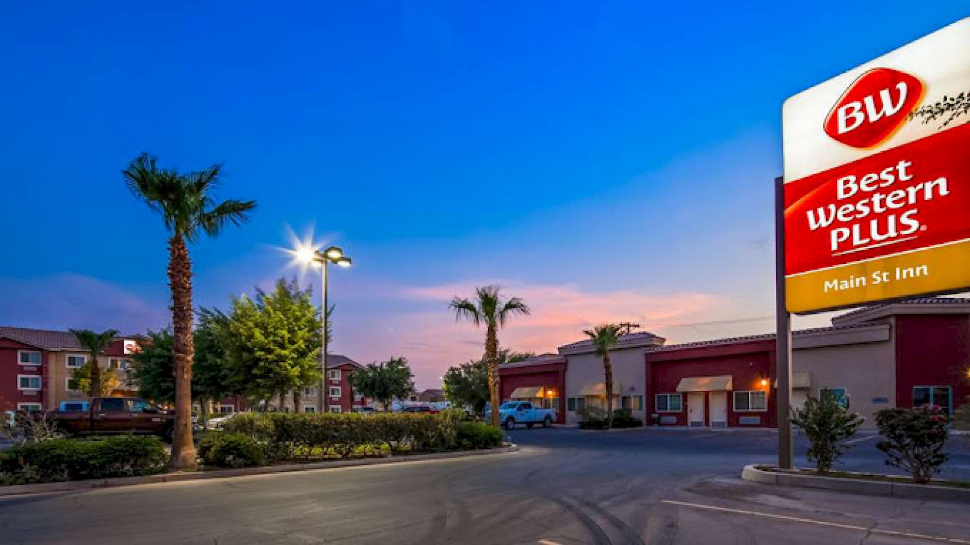 This image shows a Best Western Plus hotel with a Main St Inn sign, palm trees, and a lit parking area under a twilight sky.