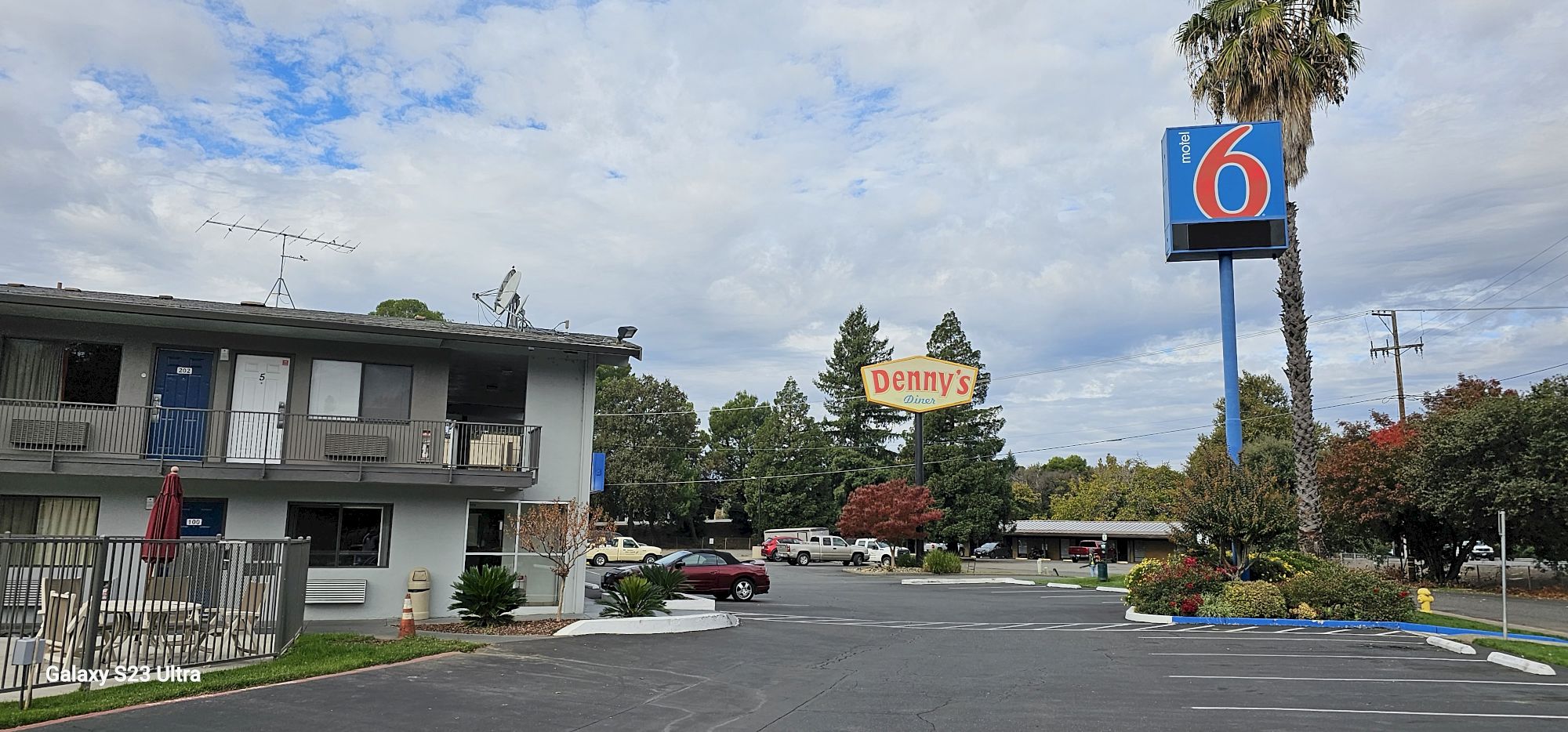 The image shows a Motel 6 parking lot with a Denny's sign nearby, surrounded by trees and a partly cloudy sky.