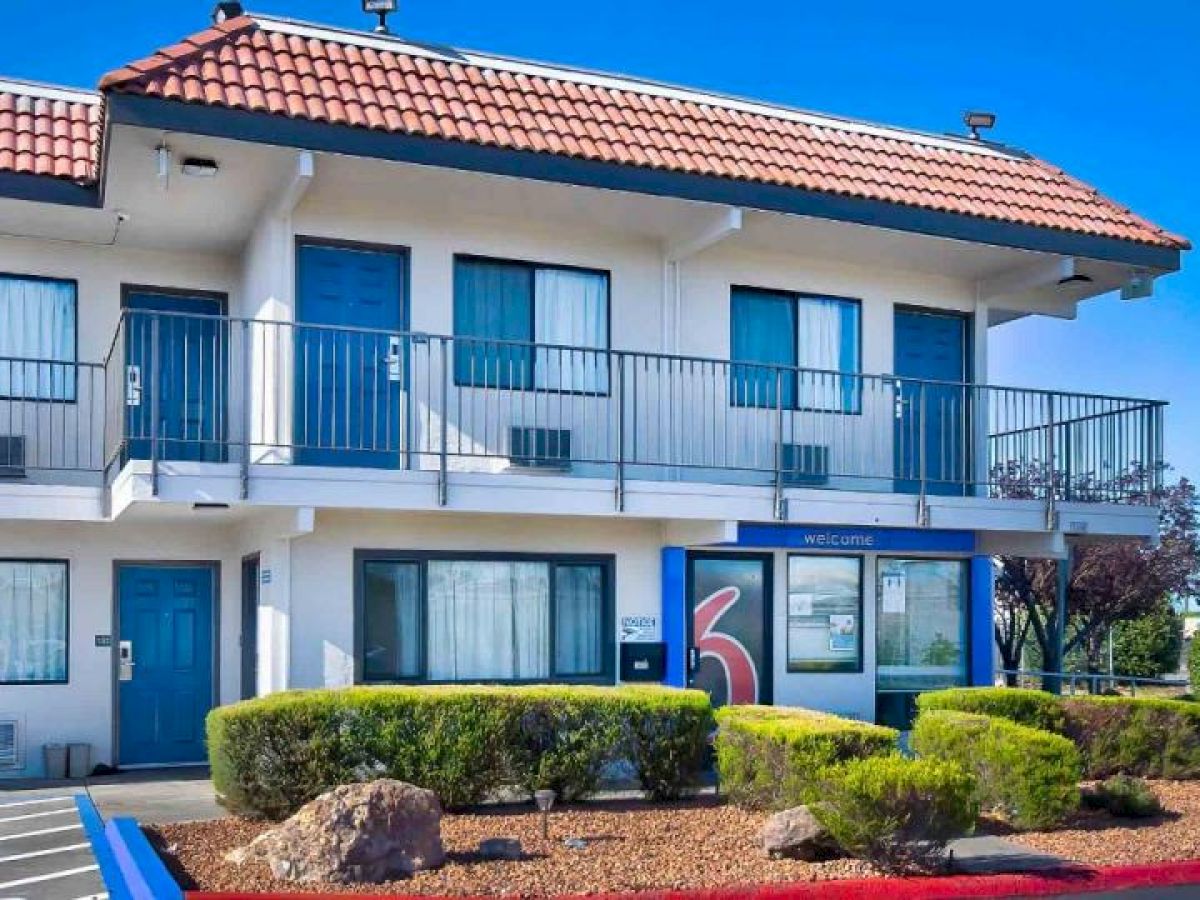 The image shows a two-story motel building with a red-tiled roof, blue doors, and manicured shrubs in front, under a clear blue sky.