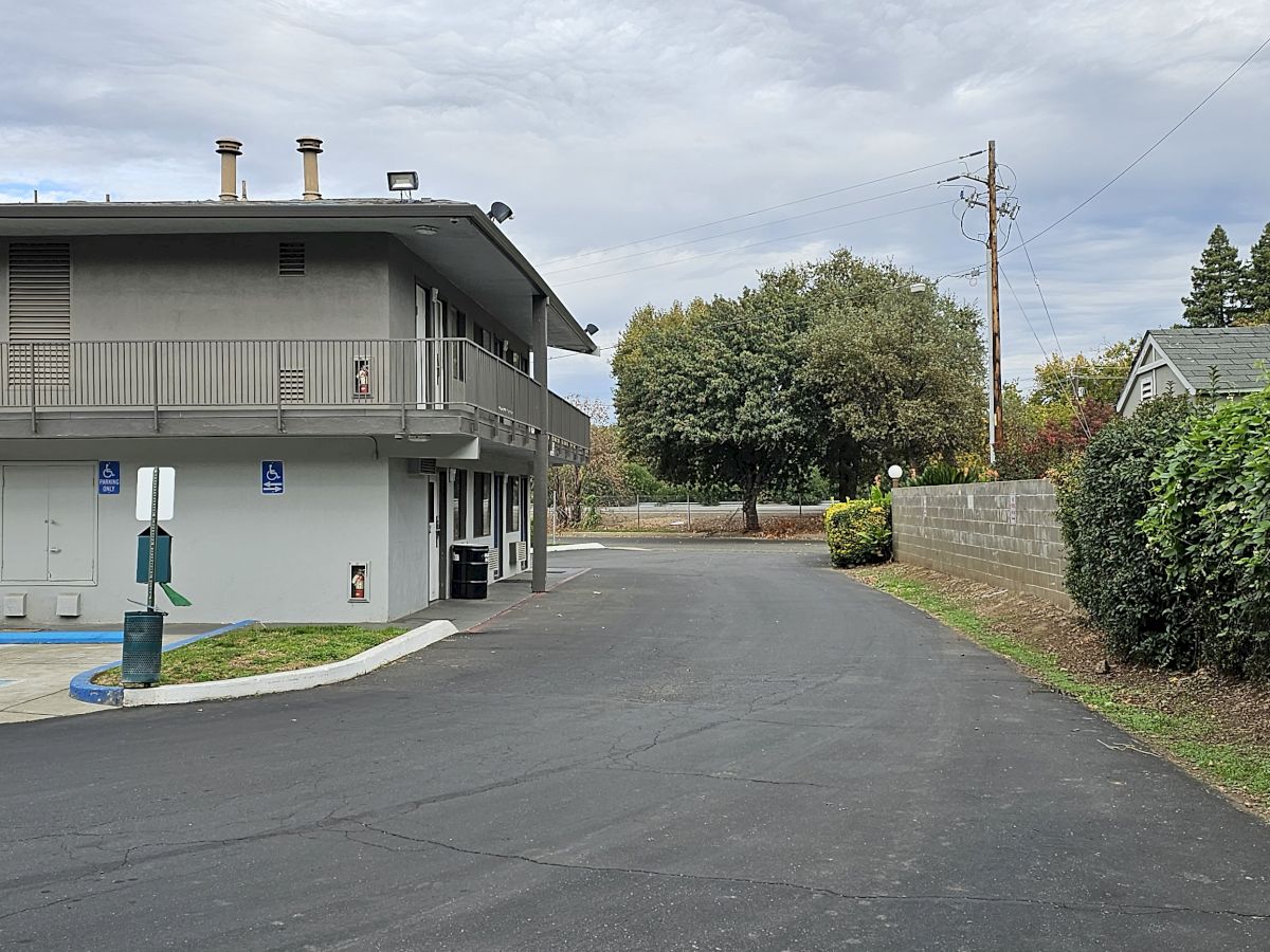 The image shows a two-story building with parking, a paved road, and greenery under a cloudy sky.