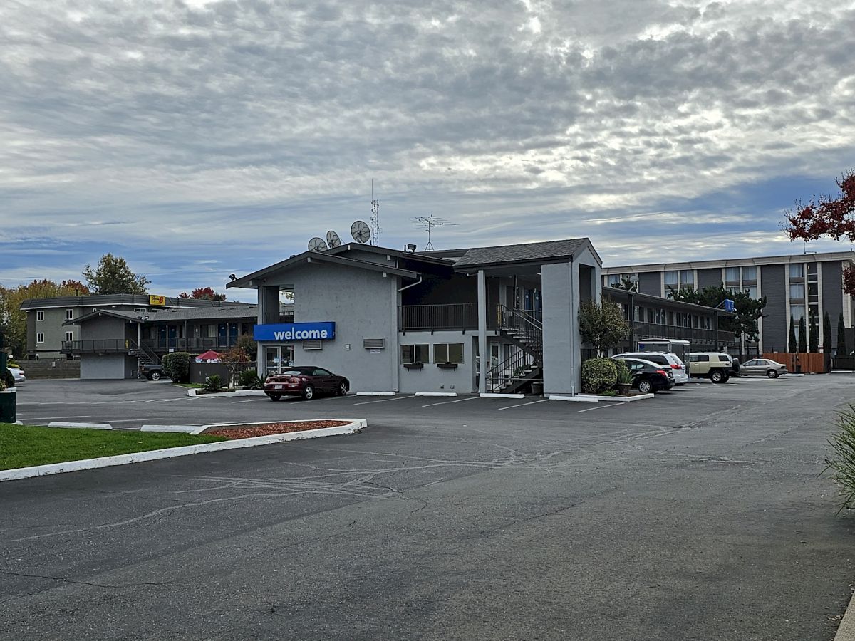 An exterior view of a Motel 6 with a parking lot and cars under a cloudy sky.