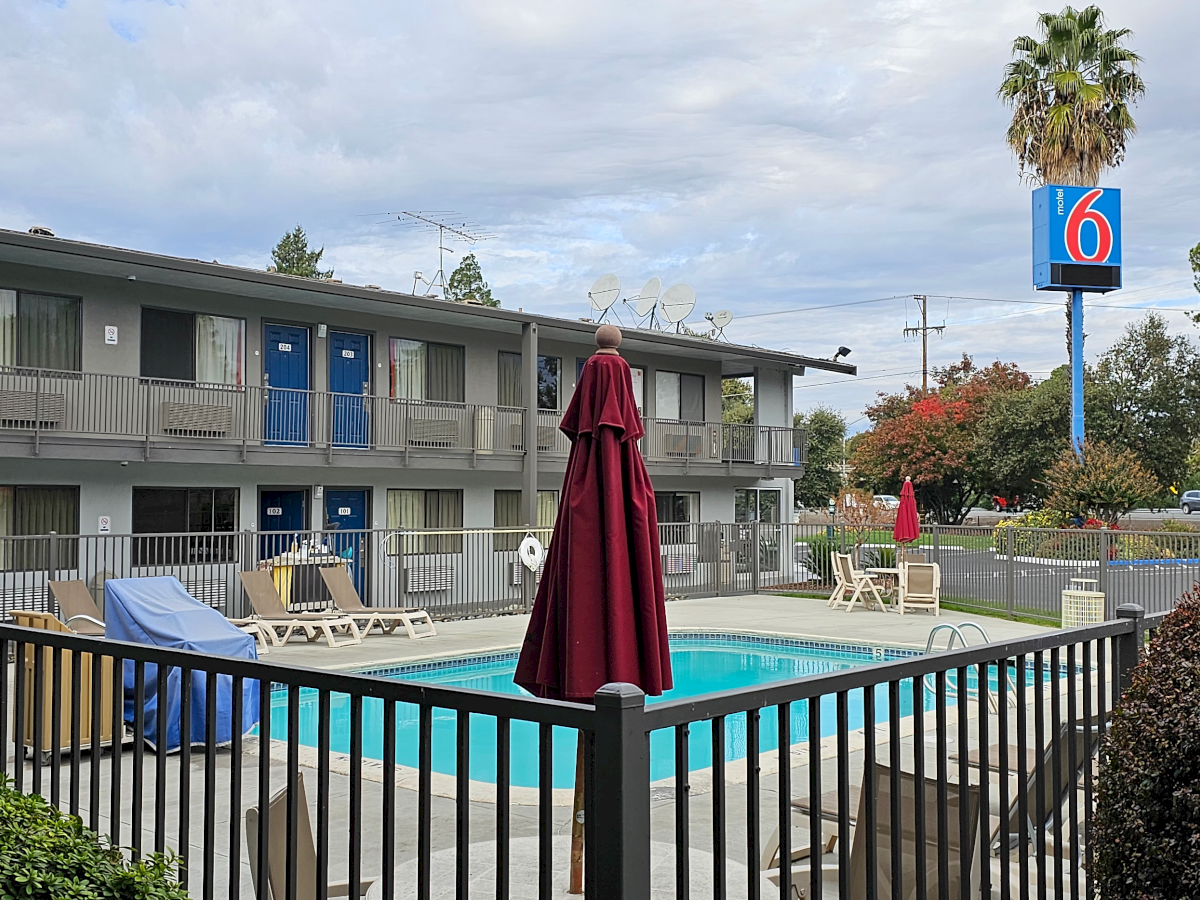 A Motel 6 with a pool, surrounded by a fence, umbrellas, and lounge chairs. The motel sign is visible amidst trees and sky.