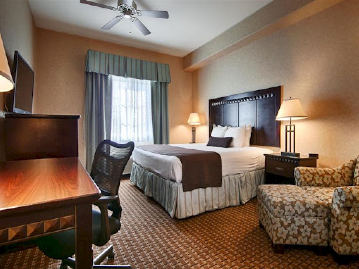 A hotel room with a bed, desk, chair, armchair, lamps, and a window with curtains, featuring warm lighting and cozy decor.