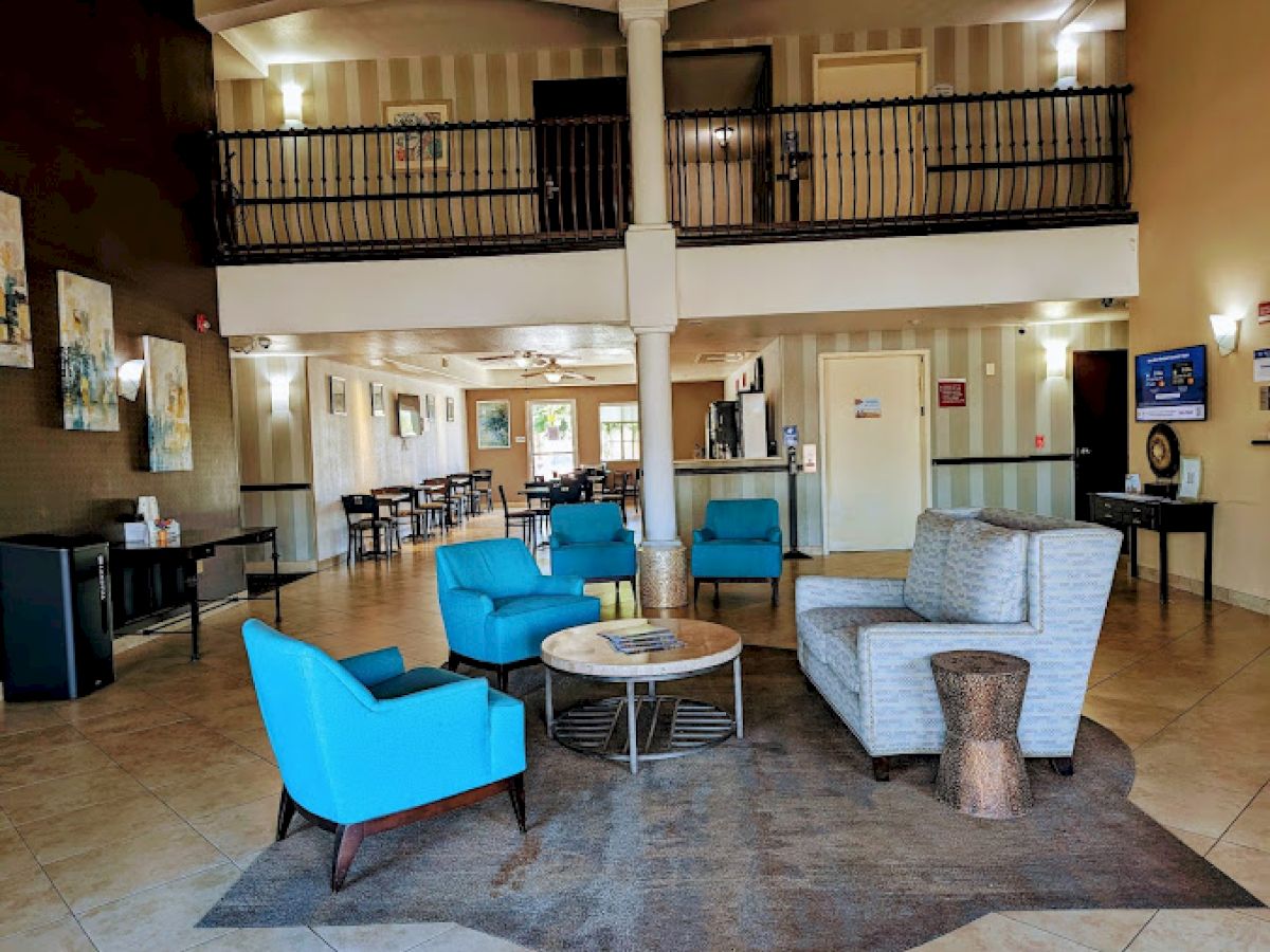 A hotel lobby with blue chairs, a gray couch, tables, and paintings. There's a balcony and a reception desk in the background.