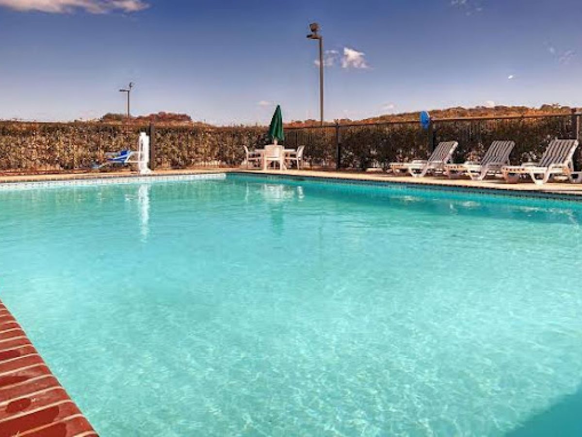 The image shows an outdoor swimming pool surrounded by lounge chairs and umbrellas, with a clear sky in the background.