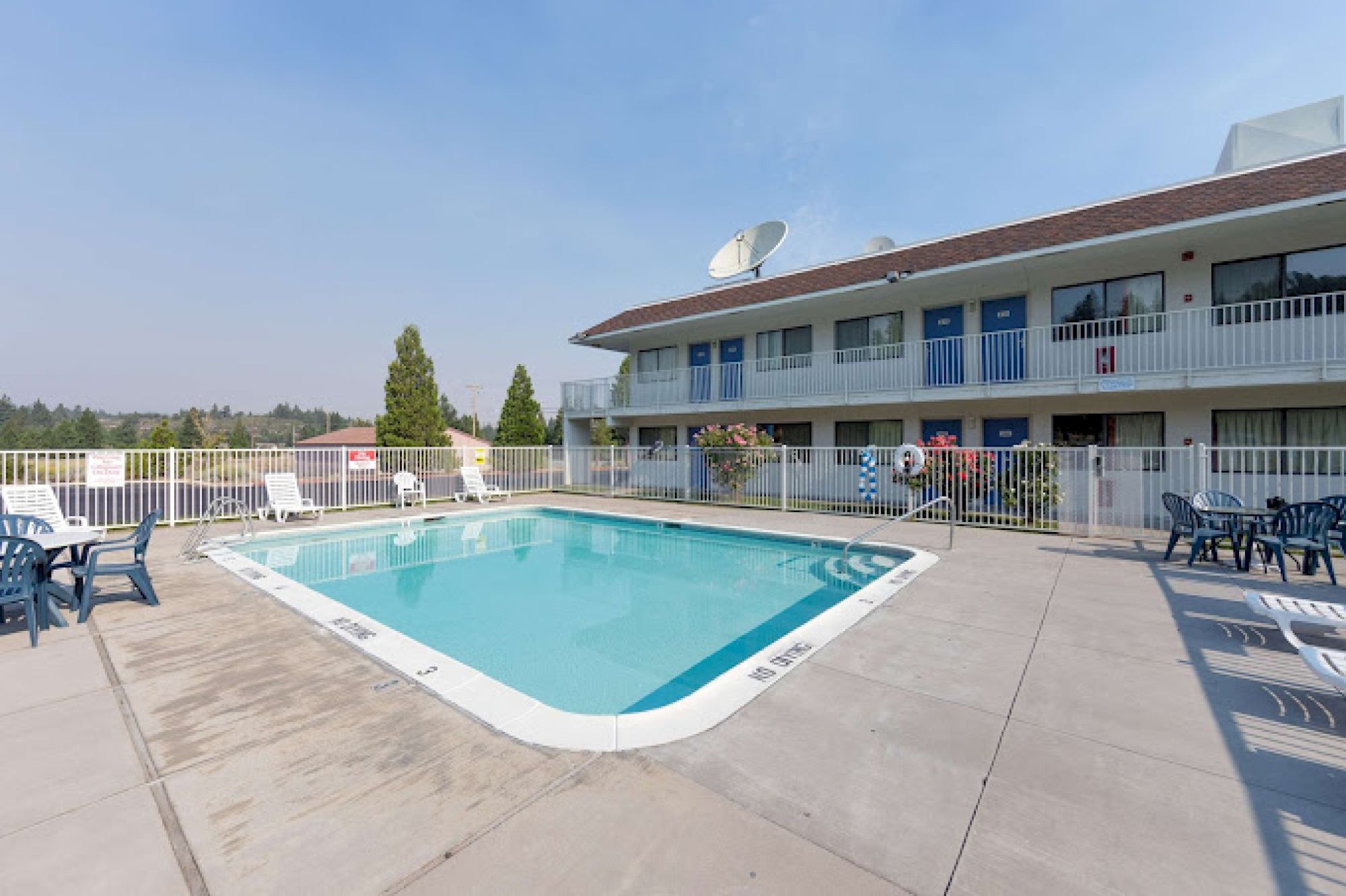 An outdoor swimming pool surrounded by a fenced area, with a two-story building and patio chairs around. Clear skies overhead complete the scene.