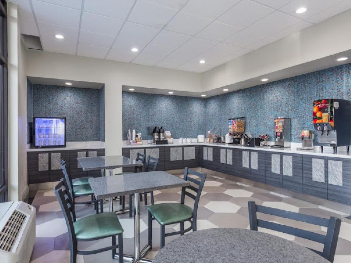 A breakfast area with tables, chairs, beverage dispensers, and a fridge. The counters are stocked with cups and condiments.