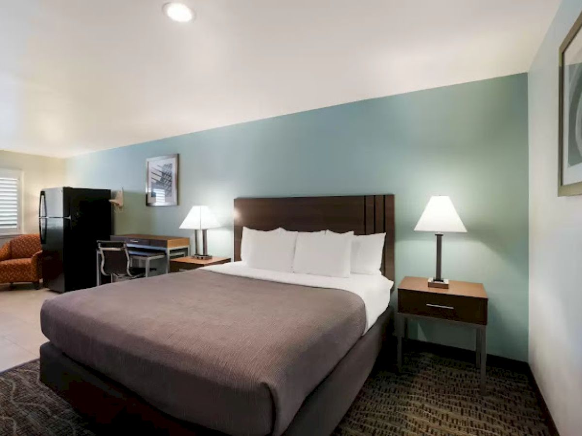 A hotel room with a bed, nightstands with lamps, a desk, a chair, and a fridge on the side. Art decorates the wall.