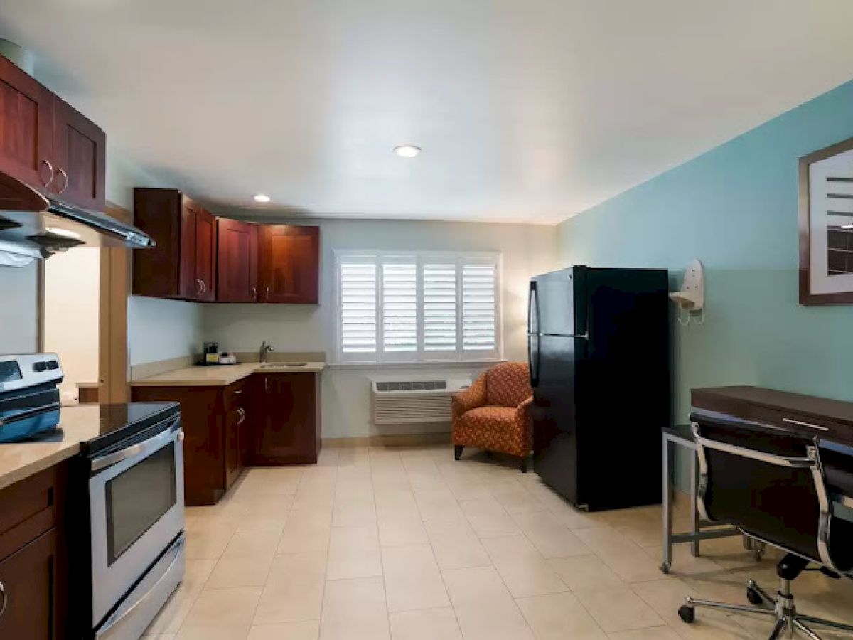 The image shows a kitchen with dark wood cabinets, an oven, a fridge, an armchair, a desk, and a window with shutters.