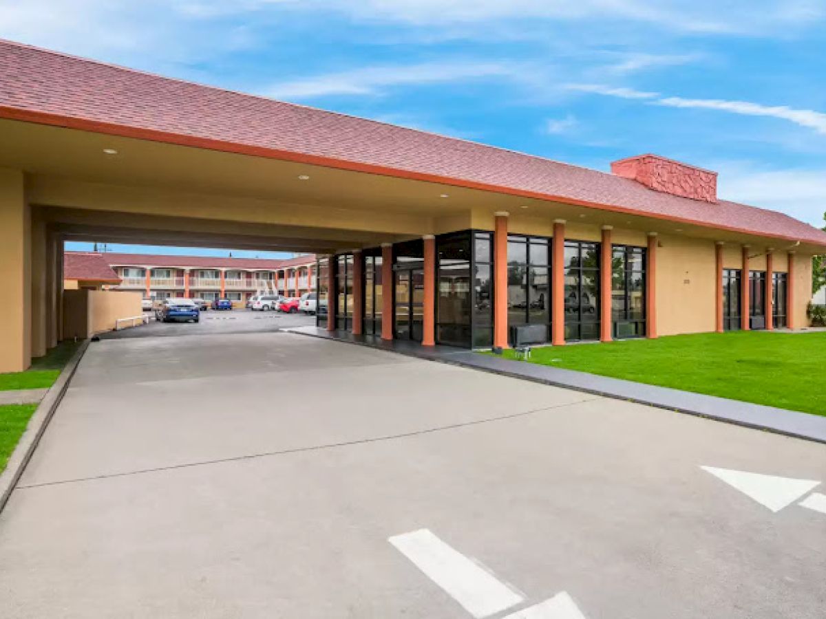 The image shows a hotel entrance with a covered driveway and glass windows, surrounded by green grass.