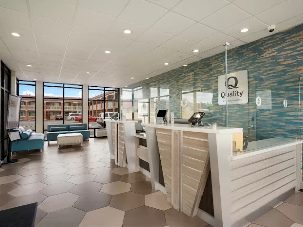 A modern hotel lobby with a front desk and sign, stylish decor, and waiting area, featuring large windows and geometric flooring.