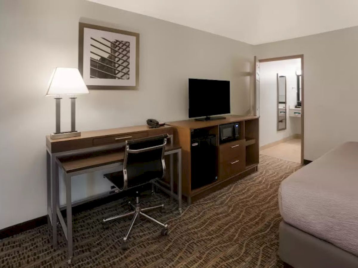 The image shows a hotel room with a desk, chair, lamp, TV, and a bed. The decor is minimal, with neutral tones throughout the space.