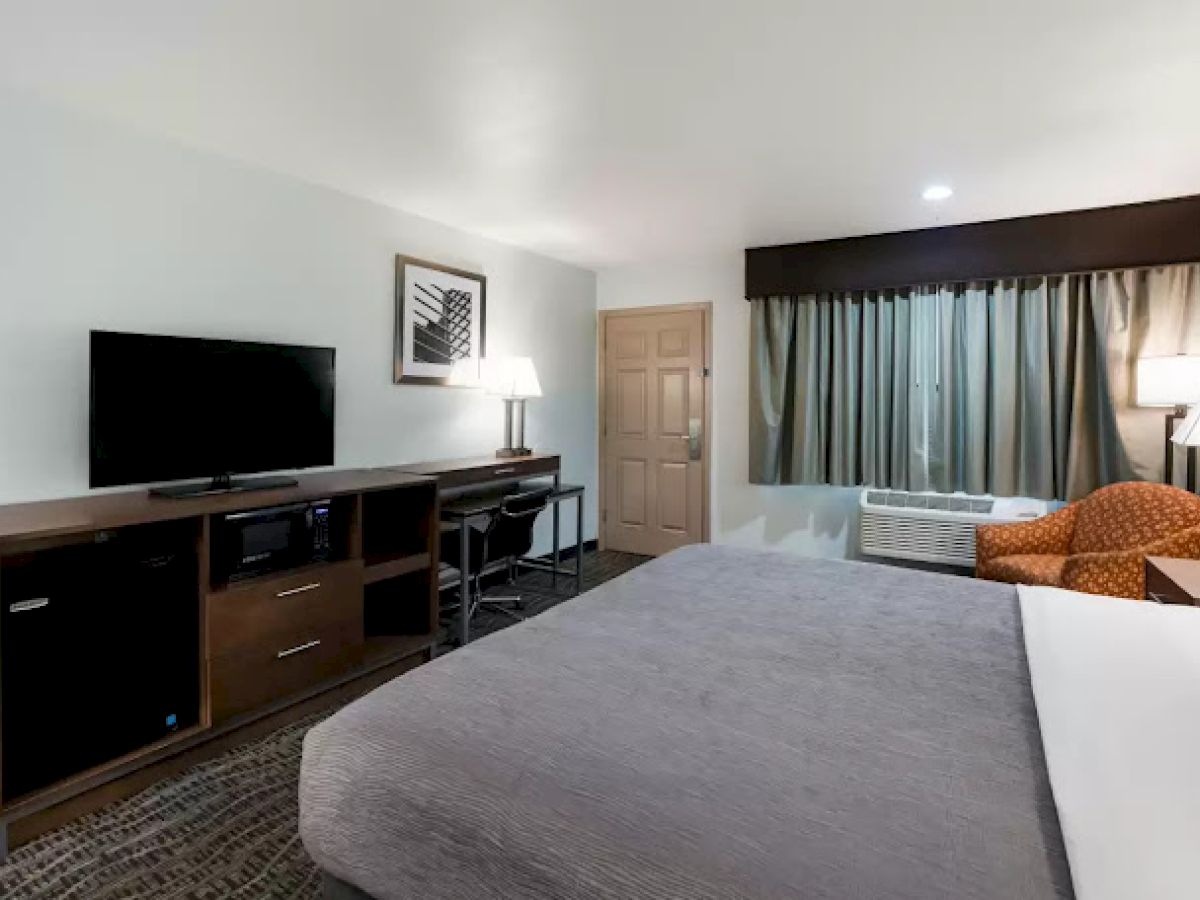 A hotel room with a bed, TV stand, desk, chair, lamps, and window with curtains. Neutral tones and simple decor complete the setting.