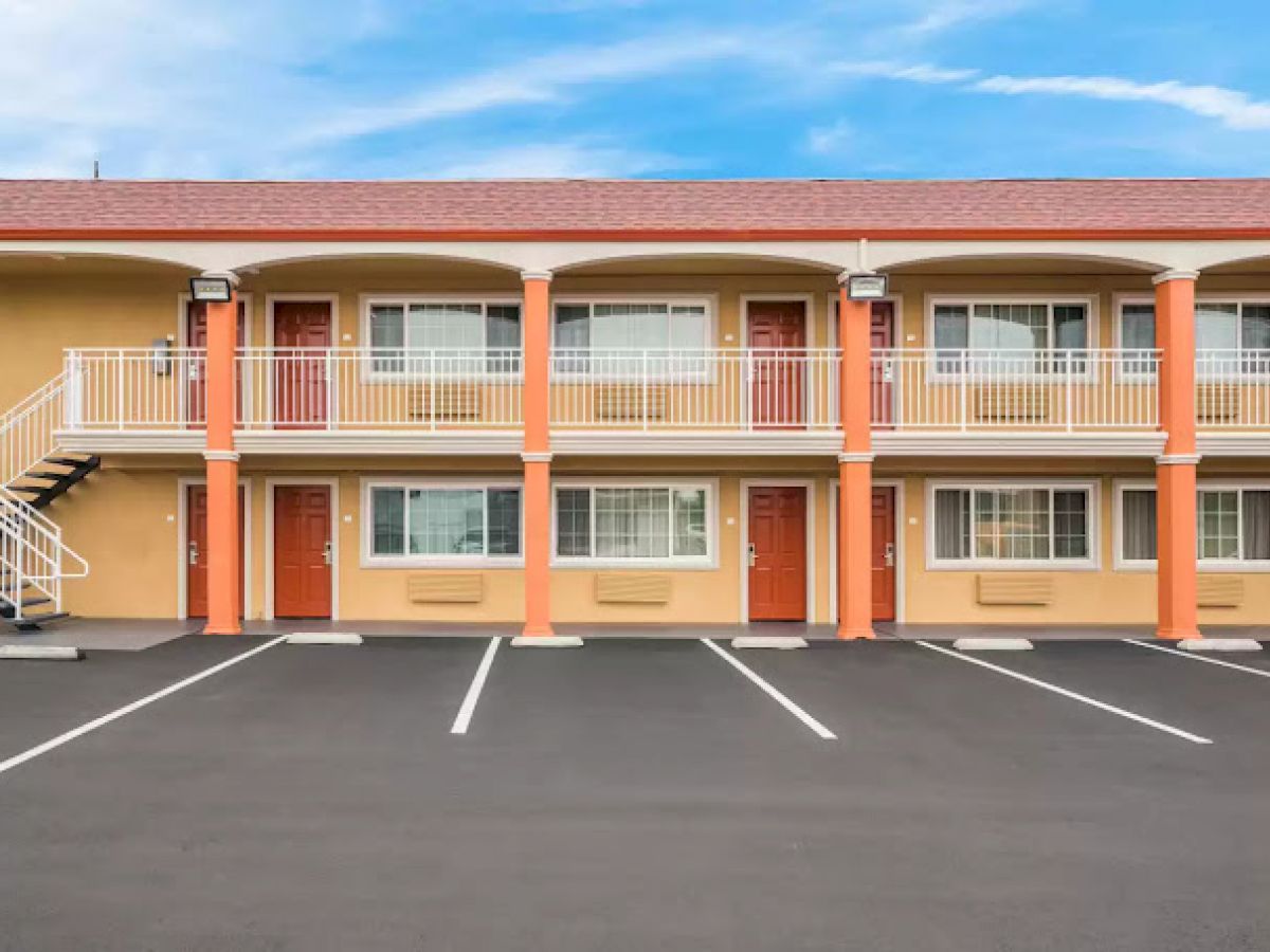 A two-story motel with orange doors and balconies is shown, featuring an empty asphalt parking lot under a clear blue sky.