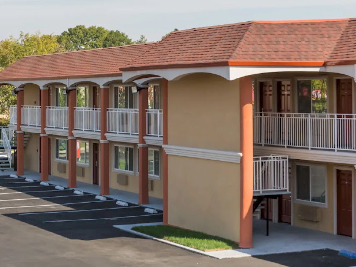 The image shows a two-story motel with an outdoor corridor, orange accents, and parked car spaces. The architectural style is simple.