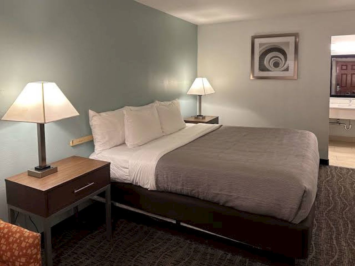 The image shows a hotel room with a bed, two lamps, nightstands, a piece of art on the wall, and a glimpse of a bathroom.