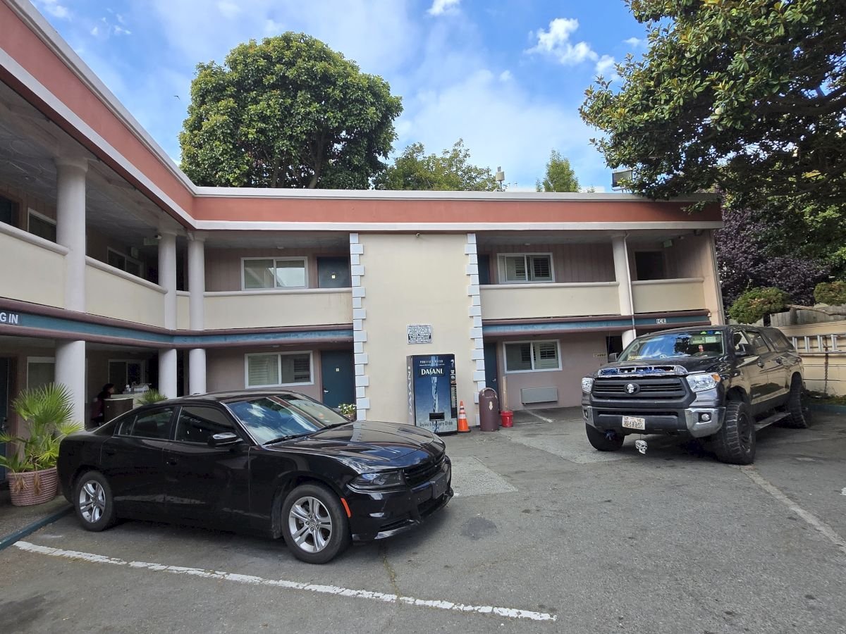 A two-story motel building with two parked cars in front and trees surrounding the area under a partly cloudy sky.