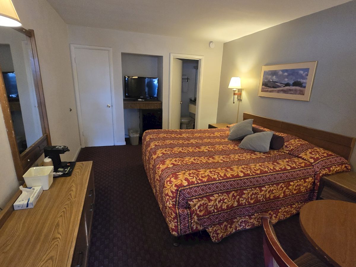 A hotel room with a large bed, TV, desk with coffee maker, and wall art. The room has a simple, cozy setup and muted lighting.