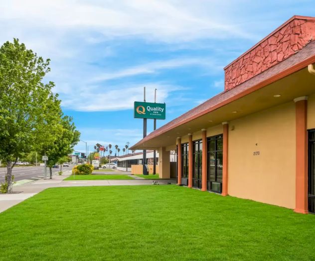 The image shows a building with a sign for Quality Inn. There's a well-maintained green lawn, a tree, sidewalk, and a blue sky.