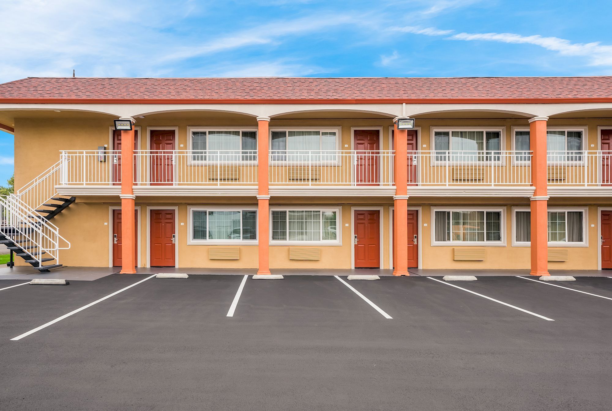 This image shows a two-story motel with red doors and a staircase, along with an empty asphalt parking lot in the foreground.