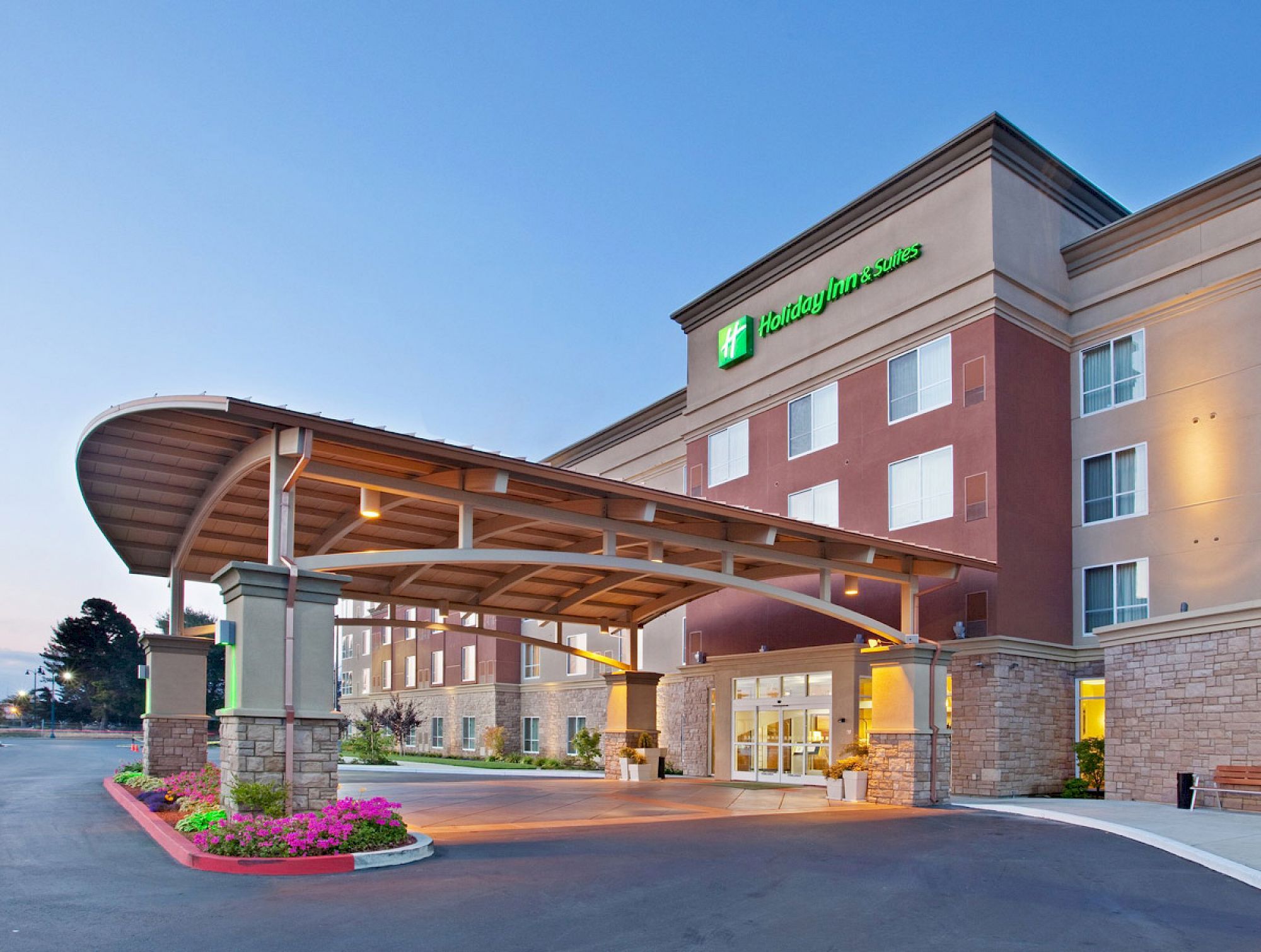 The image shows the exterior of a Holiday Inn hotel, featuring its entrance, a covered driveway, and some landscaping.