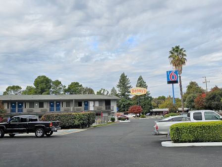 The image shows a parking lot with parked vehicles, a Motel 6 sign, and a Denny's sign, surrounded by trees and buildings under a cloudy sky.