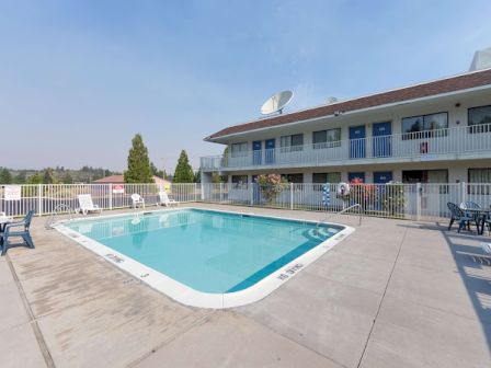 A sunny outdoor pool area with lounge chairs and tables, surrounded by a fence, next to a two-story building with blue doors and satellite dishes.