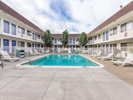 An outdoor pool surrounded by lounge chairs and a two-story motel-style building with balconies and trees in the courtyard.