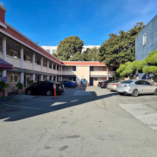 A motel courtyard with parked cars and a two-story building on a sunny day, surrounded by trees.