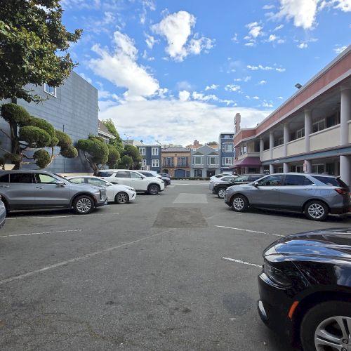 A parking lot with several cars is surrounded by buildings, under a partly cloudy blue sky.