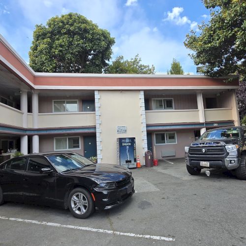 A small motel parking lot with two cars parked in front, surrounded by trees with a clear sky above.