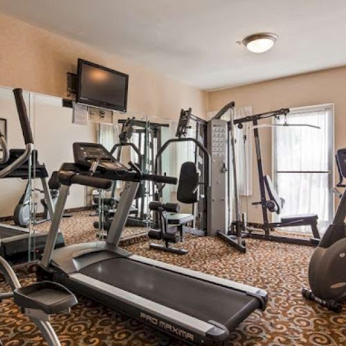 The image shows a gym room with exercise machines, including a treadmill, elliptical, and stationary bikes, with a mounted TV and clock.