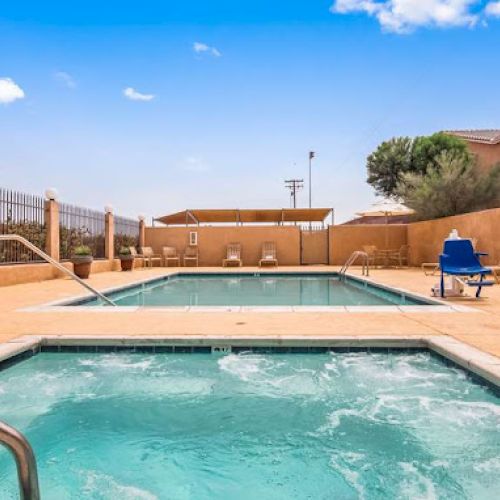 An outdoor area with a swimming pool and hot tub, surrounded by a fence, lounge chairs, blue sky, and a few buildings nearby.