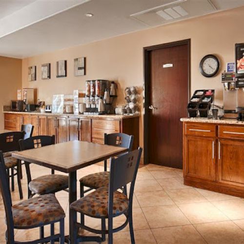 This image shows a hotel breakfast area with tables, chairs, and a countertop featuring coffee machines and breakfast supplies.