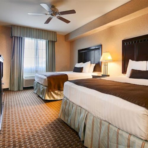 The image shows a hotel room with two beds, a TV, a window with curtains, a ceiling fan, and a lamp on a nightstand.