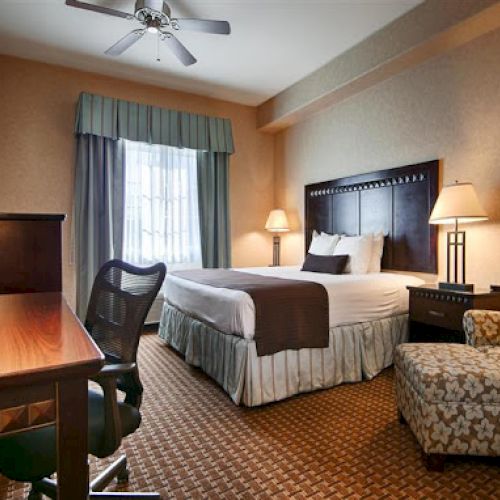 A hotel room with a large bed, desk, chairs, lamps, and patterned carpet. The room has neutral tones and a window with curtains.