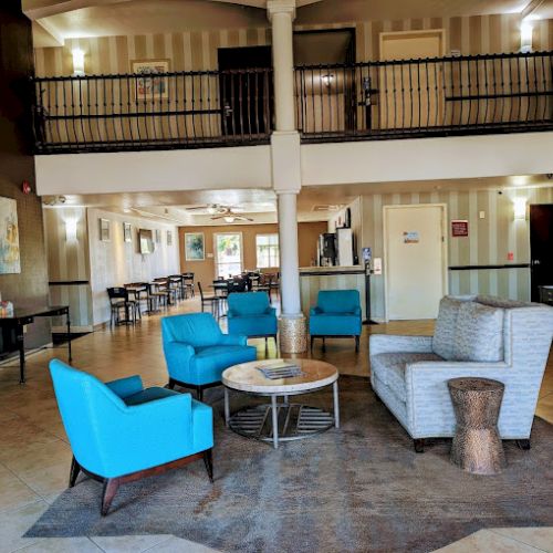 A spacious lobby with blue chairs, a gray sofa, tables, and art on the walls, featuring a staircase and upper balcony area.