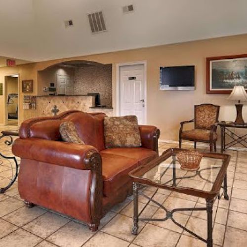 The image shows a cozy lounge area with a leather sofa, chairs, coffee table, TV, wall art, and a coffee station in the corner.